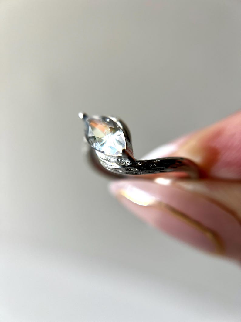 May include: A silver ring with a marquise-cut gemstone. The ring has a unique, swirling design with small accent stones. The gemstone is clear and reflects light, creating a sparkling effect. The ring is held by a person.