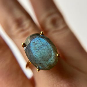 May include: A gold ring featuring a large, oval-shaped, faceted labradorite gemstone. The stone displays a prominent iridescent blue flash across its greyish surface and is secured by four small gold prongs in a classic setting design.