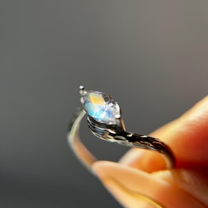 May include: A silver ring with a marquise-cut gemstone. The gemstone displays a rainbow of colors, including blue, yellow, and orange. The ring's band has a textured, organic design. The ring is held up against a gray background.
