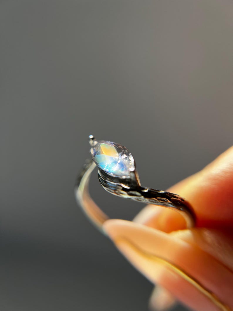 May include: A silver ring with a marquise-cut gemstone. The gemstone displays a rainbow of colors, including blue, yellow, and orange. The ring's band has a textured, organic design. The ring is held up against a gray background.