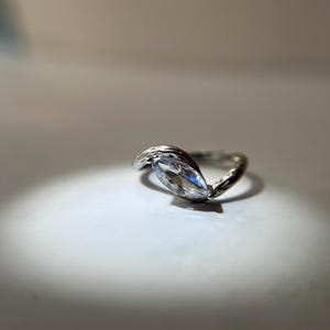 May include: A silver ring with a marquise-cut clear gemstone. The band has a textured, organic design. The ring is set against a white background, with a spotlight effect.