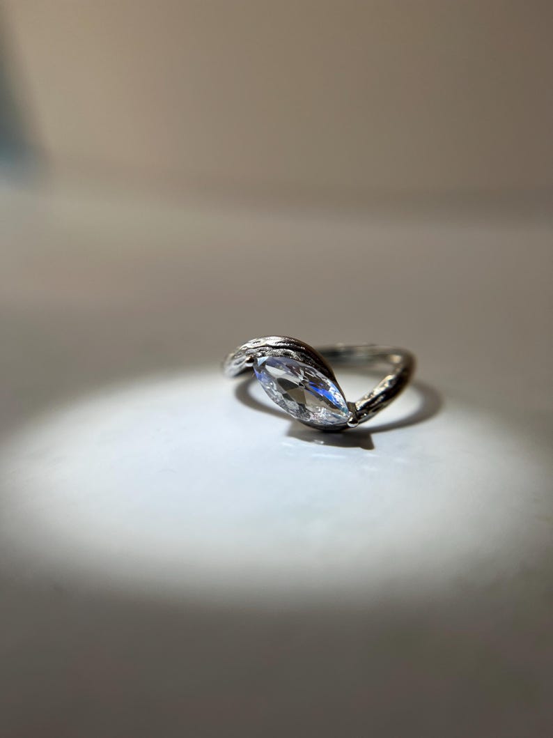 May include: A silver ring with a marquise-cut clear gemstone. The band has a textured, organic design. The ring is set against a white background, with a spotlight effect.