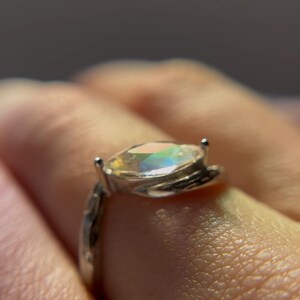 May include: A silver ring with an iridescent, oval-shaped gemstone. The ring's band has a textured design, and the gemstone reflects a spectrum of colors. A close-up shot highlights the ring's details.