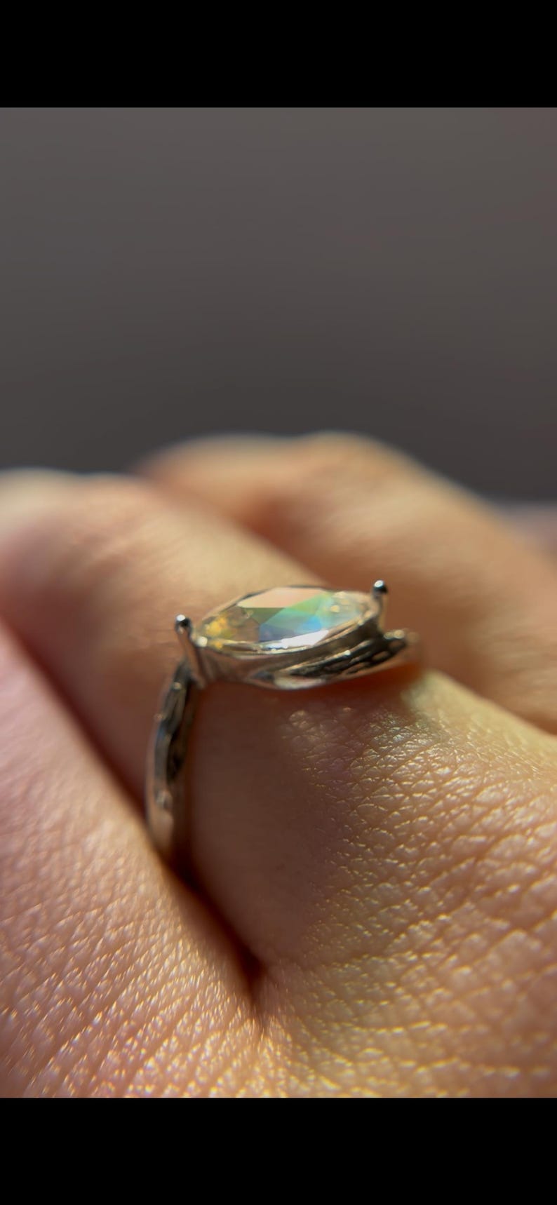 May include: A silver ring with an iridescent, oval-shaped gemstone. The ring's band has a textured design, and the gemstone reflects a spectrum of colors. A close-up shot highlights the ring's details.
