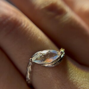 May include: Close-up of a silver ring with a marquise-cut gemstone. The gemstone has a rainbow iridescence. The ring band has a textured, organic design. The ring is on a finger.