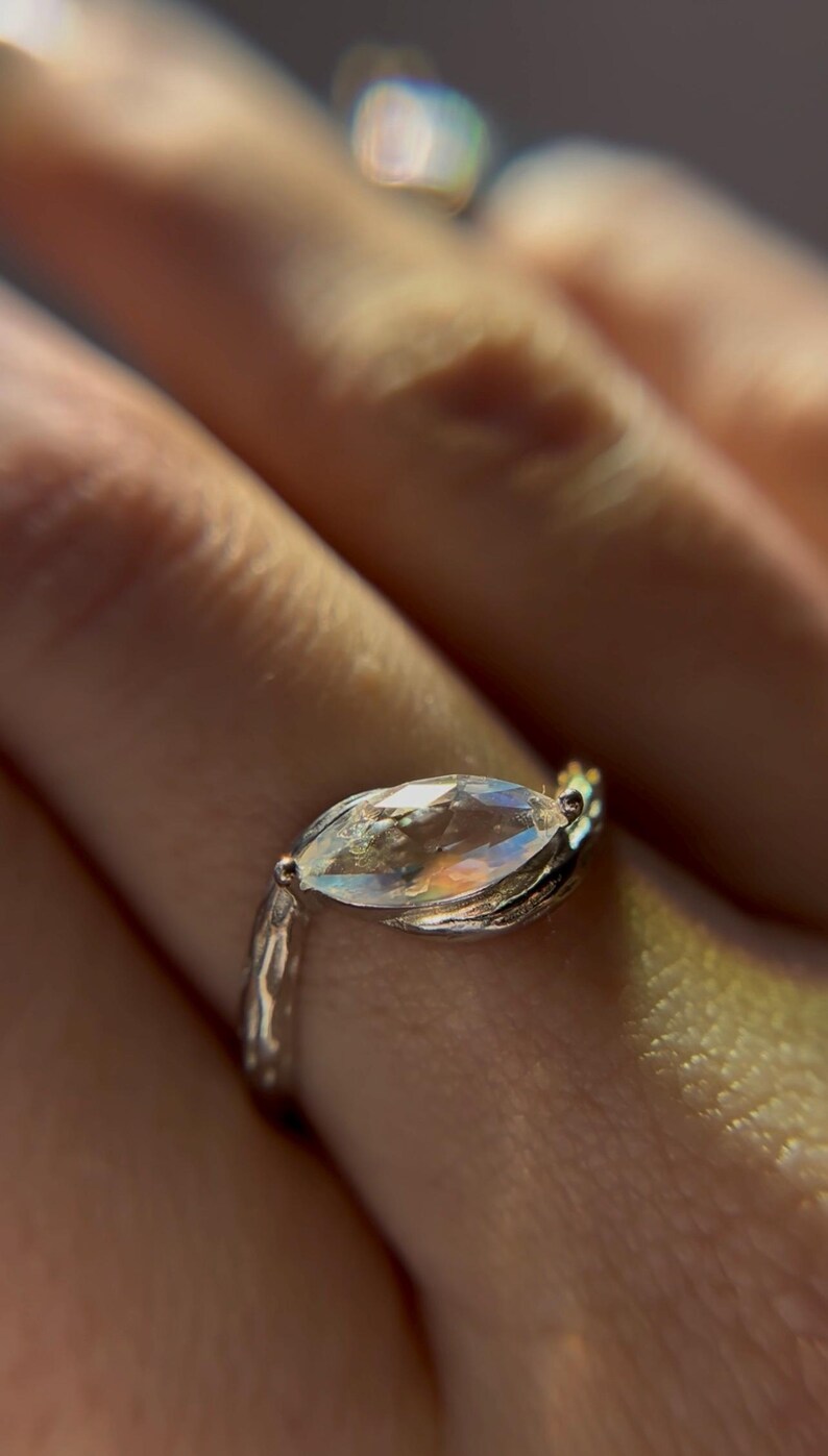 May include: Close-up of a silver ring with a marquise-cut gemstone. The gemstone has a rainbow iridescence. The ring band has a textured, organic design. The ring is on a finger.