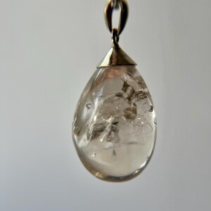 May include: A teardrop-shaped pendant made of clear, translucent quartz with visible internal inclusions and fractures. The stone is secured by a polished silver-toned metal cap and bail, which connects to a thin silver-toned chain link necklace.