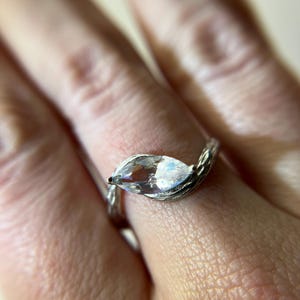 May include: A silver ring with a marquise-cut, clear gemstone. The band has a textured, twig-like design. The ring is set against a blurred background, highlighting the jewelry's details. The gemstone reflects light, creating a sparkling effect.