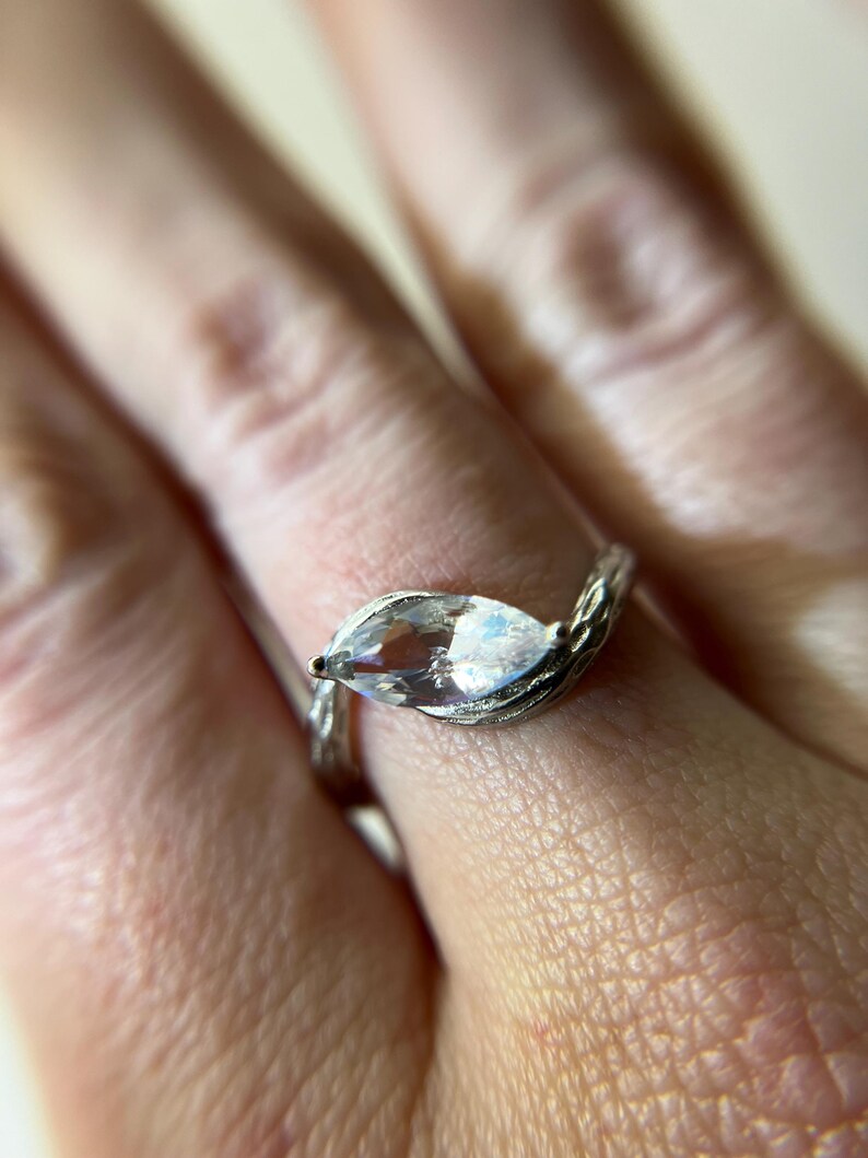 May include: A silver ring with a marquise-cut, clear gemstone. The band has a textured, twig-like design. The ring is set against a blurred background, highlighting the jewelry's details. The gemstone reflects light, creating a sparkling effect.
