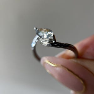 May include: A silver ring with a marquise-cut clear gemstone. The ring's band has a textured, branch-like design. The gemstone is held by a prong setting. The ring is held by a person with pink nails.