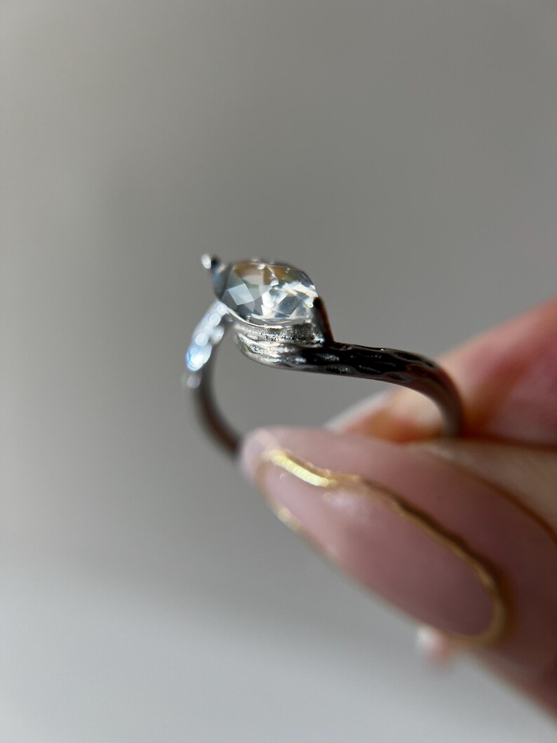 May include: A silver ring with a marquise-cut clear gemstone. The ring's band has a textured, branch-like design. The gemstone is held by a prong setting. The ring is held by a person with pink nails.