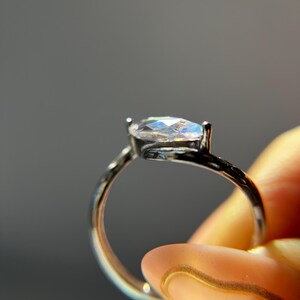 May include: A silver ring with a marquise-cut, iridescent gemstone. The ring is held up against a gray background, showcasing the gemstone's play of color. The band is thin and delicate, highlighting the central stone.