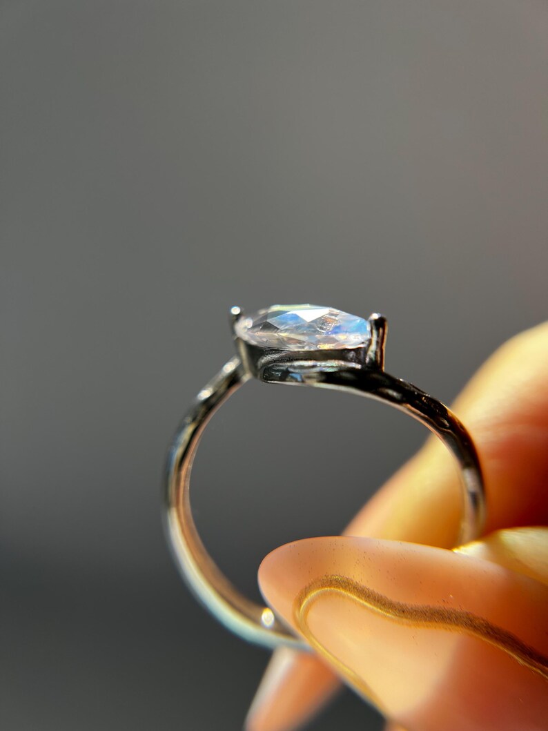 May include: A silver ring with a marquise-cut, iridescent gemstone. The ring is held up against a gray background, showcasing the gemstone's play of color. The band is thin and delicate, highlighting the central stone.