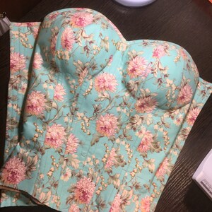 以下が含まれることがあります： ピンクとクリーム色の花柄が特徴の、ターコイズブルーのフローラルコルセット。コルセットは、ハート型のネックラインと垂直の縫い目があります。生地はキルティング加工が施されています。この衣服はおそらく手作りで、ぴったりとフィットするように設計されています。