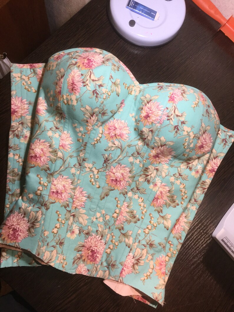 以下が含まれることがあります： ピンクとクリーム色の花柄が特徴の、ターコイズブルーのフローラルコルセット。コルセットは、ハート型のネックラインと垂直の縫い目があります。生地はキルティング加工が施されています。この衣服はおそらく手作りで、ぴったりとフィットするように設計されています。