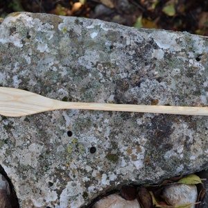 May include: A wooden spoon with a long handle and a slightly curved, rounded head. The spoon is light brown in color and is lying on a gray, textured rock.