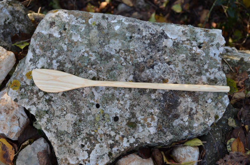 May include: A wooden spoon with a long handle and a slightly curved, rounded head. The spoon is light brown in color and is lying on a gray, textured rock.