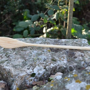 May include: A long, wooden spoon with a rounded, slightly curved head. The spoon is made of light-colored wood and is resting on a gray rock.