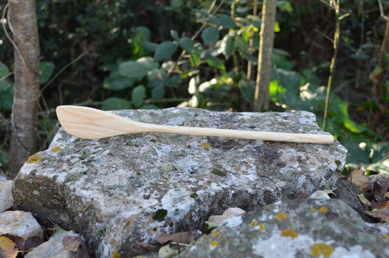 May include: A long, wooden spoon with a rounded, slightly curved head. The spoon is made of light-colored wood and is resting on a gray rock.