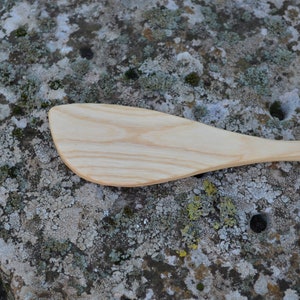 May include: A wooden spatula with a light brown color and a slightly curved shape. The spatula is lying on a gray and green textured surface.