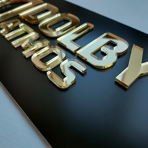 May include: A black rectangular sign with gold mirrored lettering that reads "SOLD BY". The letters are raised and have a reflective surface, creating a 3D effect. The sign is likely for a business or retail setting.
