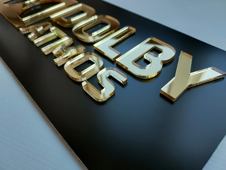May include: A black rectangular sign with gold mirrored lettering that reads "SOLD BY". The letters are raised and have a reflective surface, creating a 3D effect. The sign is likely for a business or retail setting.