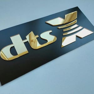May include: A black rectangular sign featuring the gold mirrored letters "dts" in a distinctive font. The letters are raised and reflective, giving a 3D appearance. The sign is likely for branding or advertising purposes.
