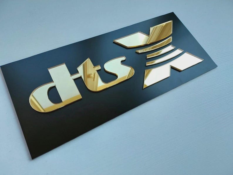 May include: A black rectangular sign featuring the gold mirrored letters "dts" in a distinctive font. The letters are raised and reflective, giving a 3D appearance. The sign is likely for branding or advertising purposes.