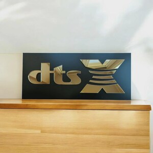 May include: A black rectangular sign featuring the gold mirrored text "dts X". The sign is placed on a wooden surface. The letters "dts" are in a stylized font, and the "X" is a geometric design.