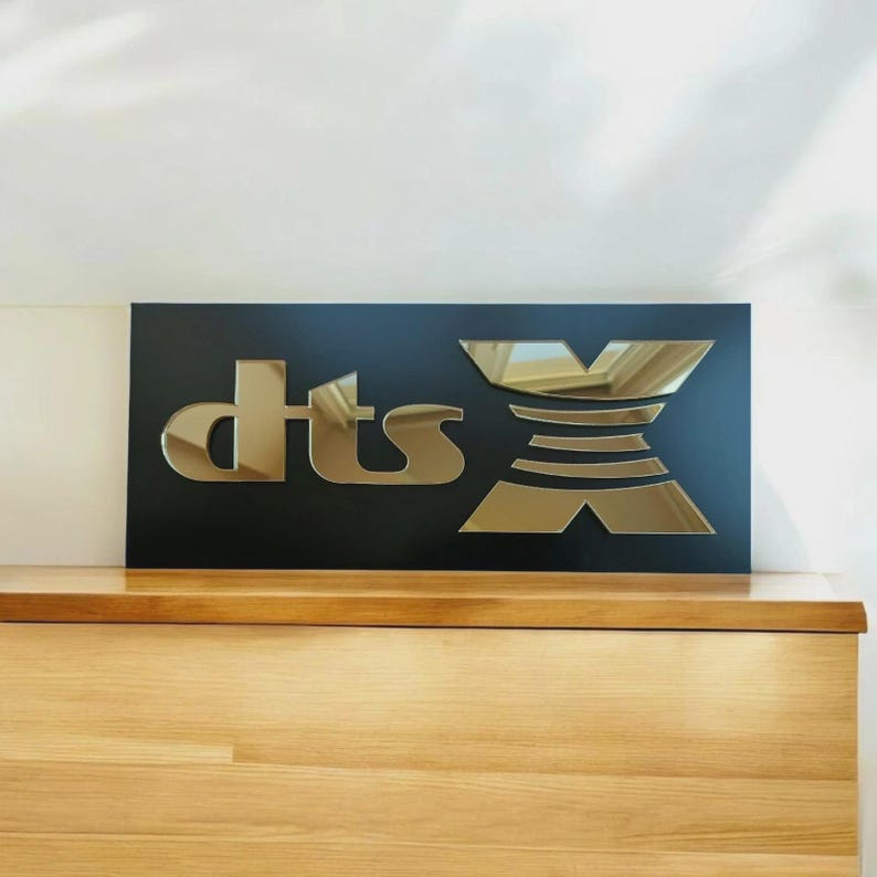May include: A black rectangular sign featuring the gold mirrored text "dts X". The sign is placed on a wooden surface. The letters "dts" are in a stylized font, and the "X" is a geometric design.