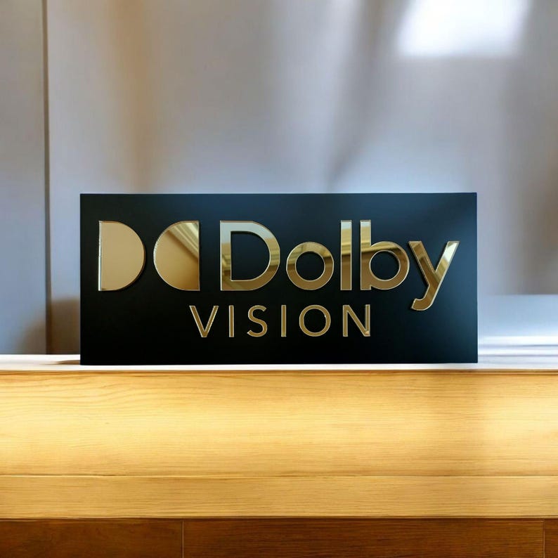 Peut inclure: Un panneau rectangulaire noir avec le texte dor&eacute; "Dolby VISION". Les lettres sont en gras, en police sans-serif. Le panneau est plac&eacute; sur une surface en bois de couleur claire.