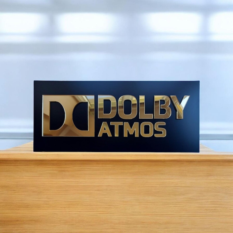 May include: A rectangular black sign with gold lettering that reads "DOLBY ATMOS." The letters are reflective and appear to be made of metal. The sign is sitting on a wooden surface.