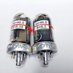Könnte beinhalten: Zwei Vintage-Vakuumröhren mit transparenten Glaskolben und silbernen Basen. Jede Röhre hat ein weißes Etikett mit schwarzem Text und ist mit einem roten Gummiband gesichert. Die Etiketten lauten "Ia=46.0" und "S=5.35" oder "S=5.3". Die Basen sind mit "MEC UV-69" gekennzeichnet.