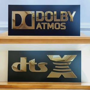 May include: Two black rectangular signs with gold lettering. The top sign reads "DOLBY ATMOS" and the bottom sign reads "dts X". The signs are displayed on a wooden surface.