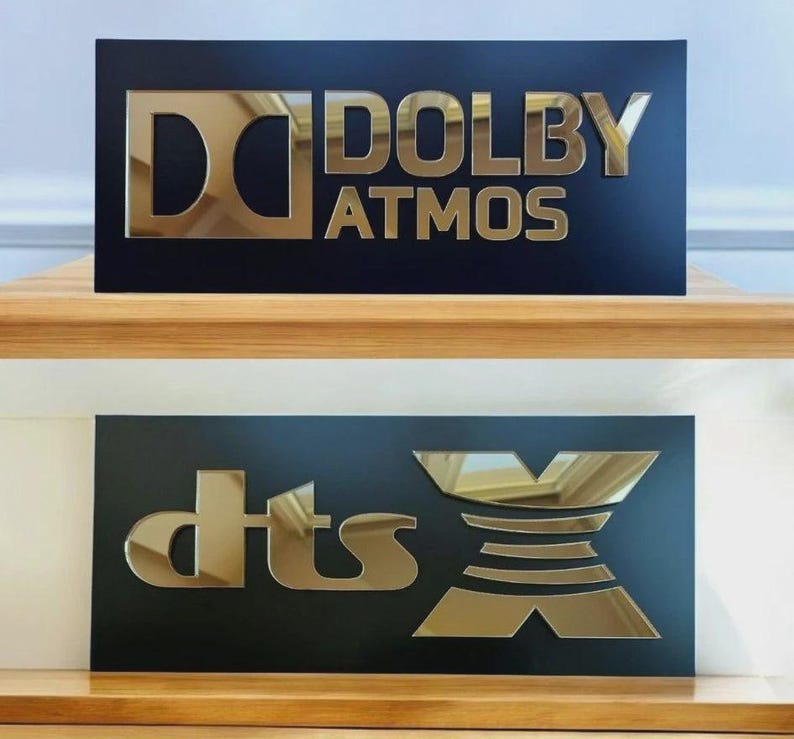 May include: Two black rectangular signs with gold lettering. The top sign reads "DOLBY ATMOS" and the bottom sign reads "dts X". The signs are displayed on a wooden surface.