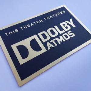May include: A rectangular sign with a gold border and a navy blue center. The sign reads "THIS THEATER FEATURES" at the top and "DOLBY ATMOS" in large, bold, gold letters. The Dolby logo is also gold.
