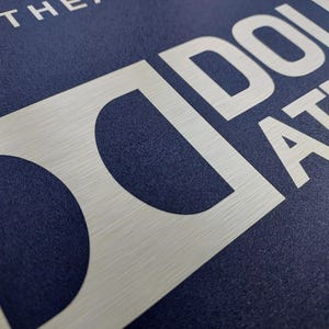 May include: Close-up of a textured navy blue surface with the word "DOLBY ATMOS" in large, brushed silver letters. The letters are bold and modern, with a slight metallic sheen.