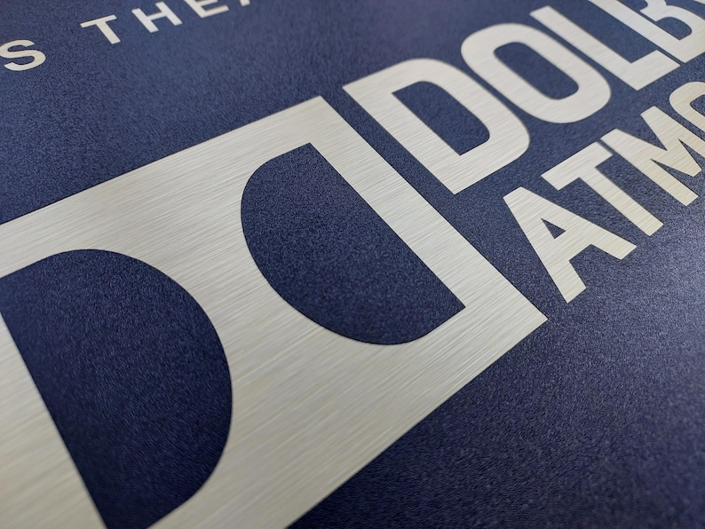May include: Close-up of a textured navy blue surface with the word "DOLBY ATMOS" in large, brushed silver letters. The letters are bold and modern, with a slight metallic sheen.