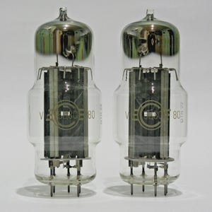 May include: Two vintage vacuum tubes with clear glass bodies and silver-coloured tops. Each tube has internal components and a base with pins. The tubes are labelled with the text "V 6230 80".