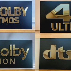 Könnte beinhalten: Vier schwarze rechteckige Schilder mit goldfarbenen, verspiegelten Buchstaben. Die Schilder zeigen die Logos für Dolby Atmos, 4K Ultra HD, Dolby Vision und DTS. Die Schilder sind wahrscheinlich für Heimkino- oder audiovisuelle Geräte.