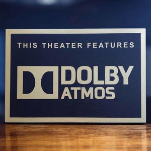May include: A rectangular sign with a gold border and navy blue center. The text "THIS THEATER FEATURES" is at the top, with "DOLBY ATMOS" in large, gold letters below. The sign is on a wooden surface.
