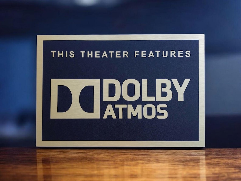 May include: A rectangular sign with a gold border and navy blue center. The text "THIS THEATER FEATURES" is at the top, with "DOLBY ATMOS" in large, gold letters below. The sign is on a wooden surface.