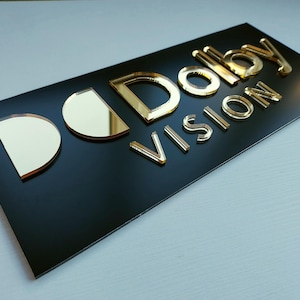Peut inclure: Panneau rectangulaire noir avec les mots "Dolby VISION" en lettres dor&eacute;es en relief. Les lettres ont un aspect m&eacute;tallique r&eacute;fl&eacute;chissant. Le panneau est pos&eacute; sur une surface claire.
