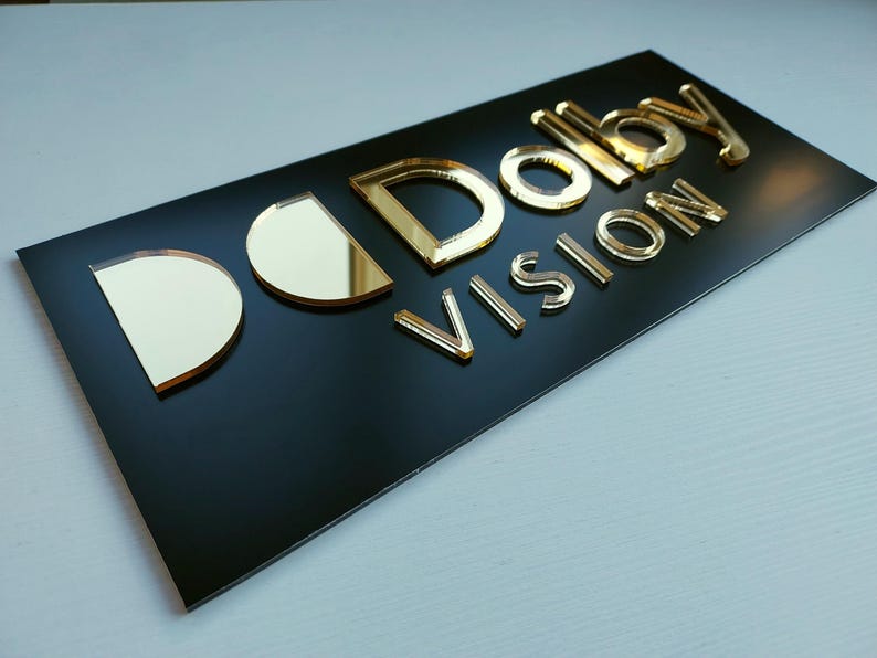 Peut inclure: Panneau rectangulaire noir avec les mots "Dolby VISION" en lettres dor&eacute;es en relief. Les lettres ont un aspect m&eacute;tallique r&eacute;fl&eacute;chissant. Le panneau est pos&eacute; sur une surface claire.