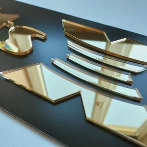 May include: Gold mirrored acrylic cutouts on a black surface. The cutouts are in various geometric shapes, reflecting the surroundings. The reflective surface contrasts with the dark background. The pieces appear to be part of a larger design, possibly a logo or decorative element.