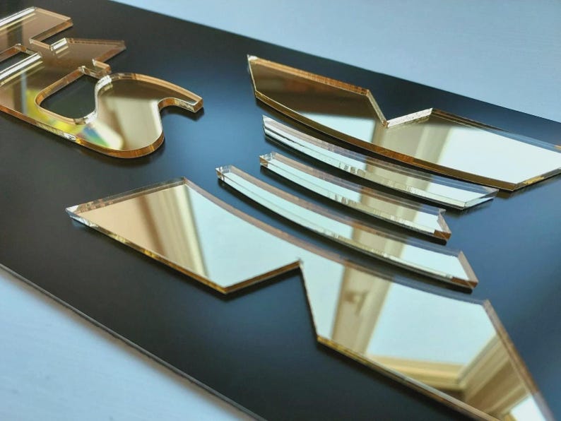 May include: Gold mirrored acrylic cutouts on a black surface. The cutouts are in various geometric shapes, reflecting the surroundings. The reflective surface contrasts with the dark background. The pieces appear to be part of a larger design, possibly a logo or decorative element.