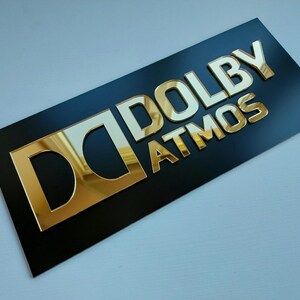 May include: A black rectangular sign with the gold text "DOLBY ATMOS". The letters are raised and have a reflective, metallic appearance. The "D" logo is also gold.