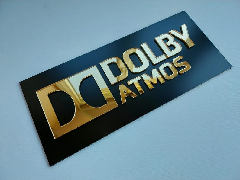 May include: A black rectangular sign with the gold text "DOLBY ATMOS". The letters are raised and have a reflective, metallic appearance. The "D" logo is also gold.