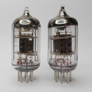 May include: Two vintage vacuum tubes with glass enclosures and metal bases. The tubes have multiple pins and are labelled "VII 1953" and "VII 1953".
