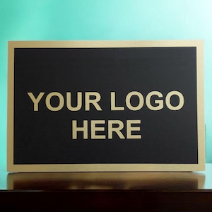 May include: A rectangular sign with a black centre and a gold border. The words "YOUR LOGO HERE" are printed in gold, centred on the black background. The sign is displayed on a dark brown surface.